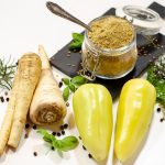 A flat lay of parsnips, bell peppers, herbs, and a spice jar on a white background.