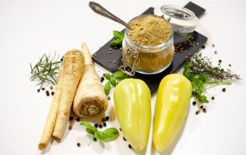 A flat lay of parsnips, bell peppers, herbs, and a spice jar on a white background.